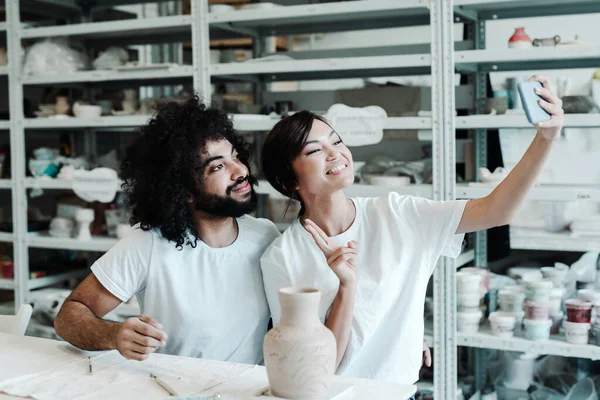 Un hombre y una mujer enamorados se toman un selfie en un taller de cerámica, sentados en una mesa con una olla de barro y pinceles para pintar. Citas en un estudio de hobby, pasar el tiempo libre Fotos De Stock