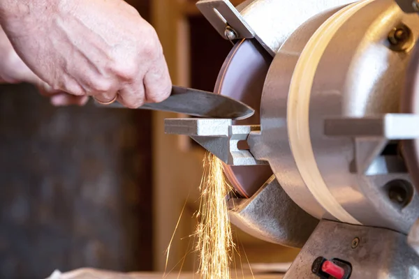 Sharpening a knife on an electric grinder close-up. Male hands hold the knife at the sharpening machine, bright sparks fly off the rotating disc.