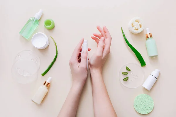 Mujer que aplica crema orgánica en las manos, hojas de aloe vera y productos cosméticos alrededor de la superficie beige. Vista superior — Foto de Stock