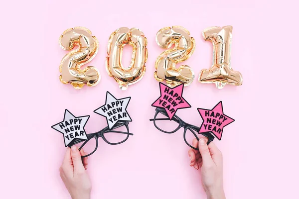 Balony foliowe w postaci cyfr 2021 i świąteczne okulary w rękach kobiet. Święto Nowego Roku. Widok z góry na różowe tło — Zdjęcie stockowe