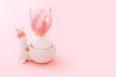 Paskalya yumurtası seramik Paskalya tavşanı heykelciği ve pastel pembe arka planda bahar lale çiçeğiyle duruyor.