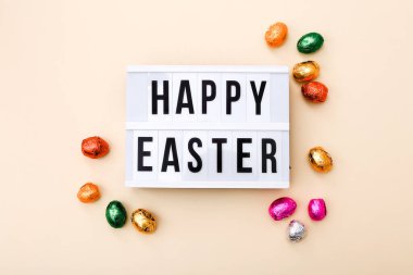 Paskalya dilekleri, bej arka planda renkli bir folyoya sarılı çikolatalı mini yumurtalarla ışık kutusunda.