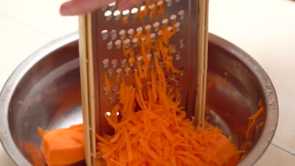 Las manos femeninas rallan firmemente la zanahoria en el rallador metálico y en el bol. Zanahorias ralladas en un tazón rodeado de verduras. Preparación de comida vegana — Vídeo de stock