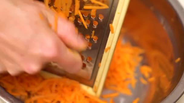 Las manos femeninas rallan firmemente la zanahoria en el rallador metálico y en el bol. Zanahorias ralladas en un tazón rodeado de verduras. Preparación de comida vegana — Vídeo de stock