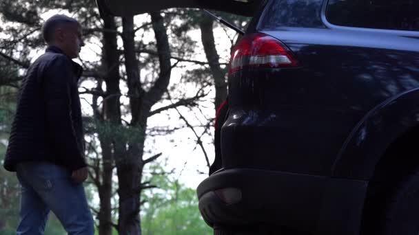 Az ember kinyitja csomagtartó egy SUV a természetben, és húzza ki a turisztikai asztal és szék — Stock videók