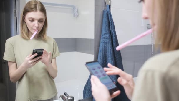 A young woman brushes her teeth. Phone in hand. — Stock Video