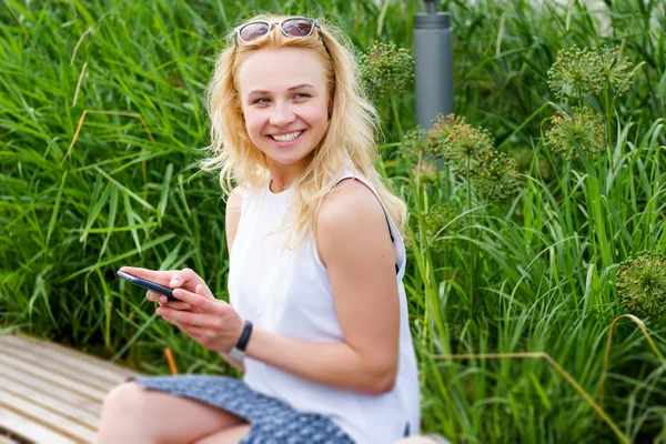 Кавказская девушка сидит на скамейке в парке и держит телефон — стоковое фото