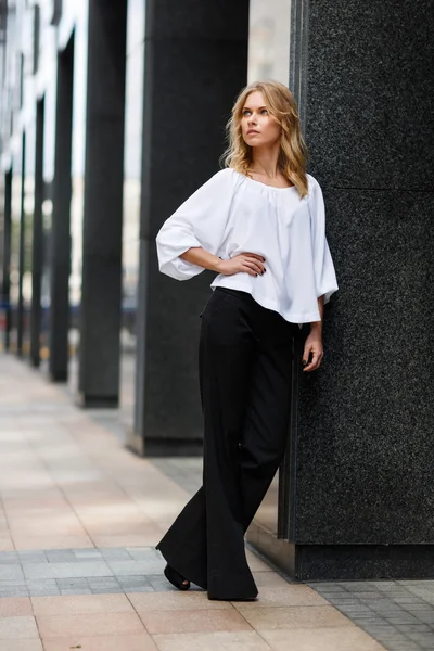 Блондинка в белой блузке и черных брюках возле здания — стоковое фото