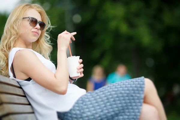 Hermosa chica rubia sentada en el banco con una taza de bebida — Foto de Stock