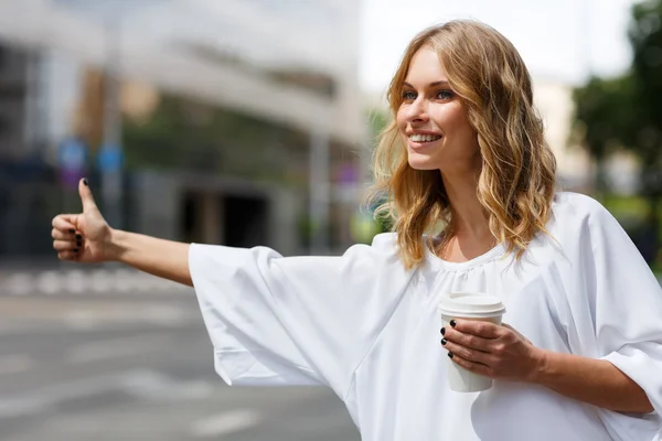Hübsche Frau hält Kaffee und stoppt Transport mit erhobenem Daumen — Stockfoto