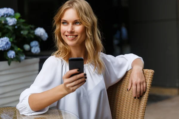 Улыбающаяся блондинка в белой блузке с телефоном в руке — стоковое фото