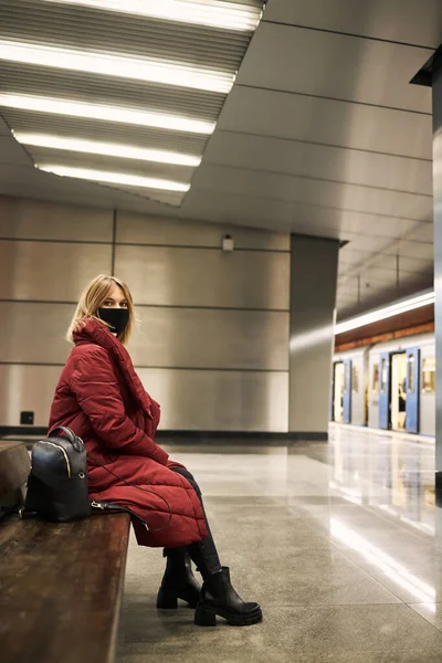 Una chica en una máscara se sienta y espera un tren — Foto de Stock