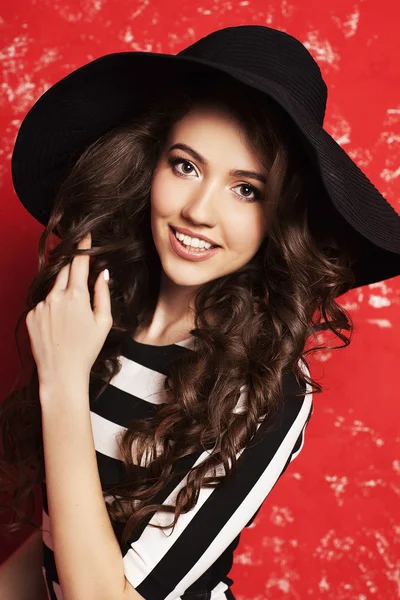 Portrait of beautiful young woman with long curly hair in black hat and striped dress on red background. — Stock Photo, Image