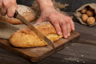 Ekmek bıçağıyla erkek elleri tahta bir tahtanın üzerinde taze taze kesilmiş baget ekmeği. Dilimlenmiş geleneksel Fransız ekmeği. Yakın plan.