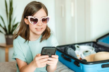 Genç gezgin akıllı telefondan gezi planlıyor. Evde bavul ve gözlüklü çocuk.