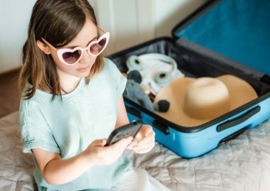 Genç gezgin akıllı telefondan gezi planlıyor. Evde bavul ve gözlüklü çocuk.