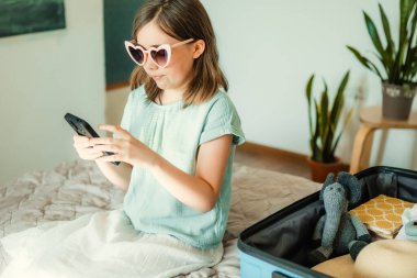 Genç gezgin akıllı telefondan gezi planlıyor. Evde bavul ve gözlüklü çocuk.