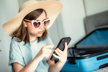 Küçük tatlı bir kız yaz gezisi için hazırlanıyor. Yatak odasında bir yatakta oturuyor, şık bir hasır şapka ve kalp şeklinde güneş gözlüğü takıyor. Yanında içi boş mavi bir bavul var.