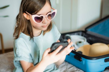 Genç gezgin akıllı telefondan gezi planlıyor. Evde bavul ve gözlüklü çocuk.