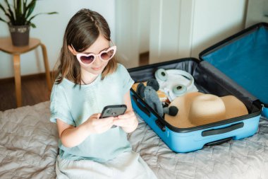 Genç gezgin akıllı telefondan gezi planlıyor. Evde bavul ve gözlüklü çocuk.