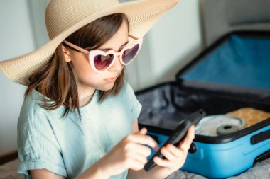 Küçük tatlı bir kız yaz gezisi için hazırlanıyor. Yatak odasında bir yatakta oturuyor, şık bir hasır şapka ve kalp şeklinde güneş gözlüğü takıyor. Yanında içi boş mavi bir bavul var.