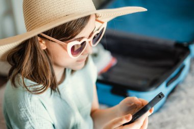 Küçük tatlı bir kız yaz gezisi için hazırlanıyor. Yatak odasında bir yatakta oturuyor, şık bir hasır şapka ve kalp şeklinde güneş gözlüğü takıyor. Yanında içi boş mavi bir bavul var.