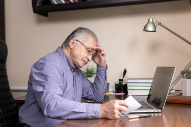 Kıdemli adam bilgisayar kullanmayı öğrendi. Cam ve mavi gömlekli yaşlı adam ev ofisinde online çalışmak için dizüstü bilgisayar kullanma talimatlarını okuyor.