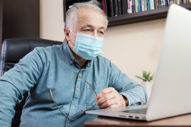 Maskeli kıdemli adam evde bilgisayarla çalışıyor ya da iletişim kuruyor. Coronavirus döneminde eğitim, eğitim, iş, iletişim, eğlence, boş zaman.