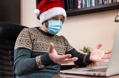 Noel online tebrikleri. Noel Baba şapkalı ve maskeli genç adam bilgisayar kullanarak arkadaşları ve çocukları arayarak şok etti ve şaşırttı. Coronavirus sırasında Noel.
