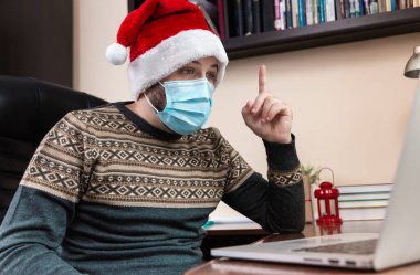 Noel online tebrik fikri. Noel Baba şapkalı ve maskeli genç adam bilgisayar kullanarak arkadaşlarını ve çocuklarını arıyor. Coronavirus sırasında Noel.