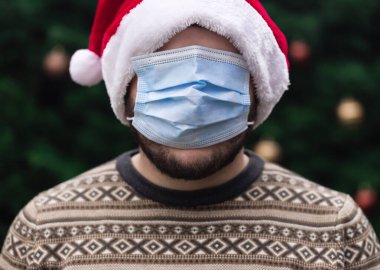 Maskeyi yanlış takıyorsun. Noel Baba şapkası takan bir adamın portresi, Xmas kazağı ve duygu yüklü tıbbi maske. Bir Noel ağacının arka planında. Koronavirüs salgını