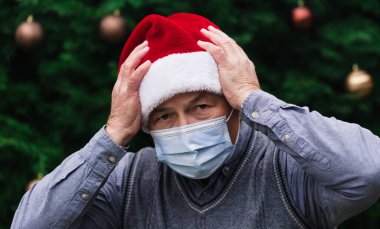 Şaşırmış ya da şaşırmış bir Noel. Noel Baba şapkası ve tıbbi maske takan son sınıf öğrencisinin portresi. Bir Noel ağacının arka planında. Koronavirüs salgını