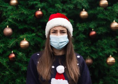 Noel Baba şapkası takan ve tıbbi maske takan kadın portresi. Bir Noel ağacının arka planında. Koronavirüs salgını