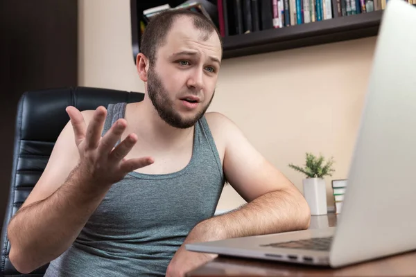 Leisure using laptop. A young bearded guy in home clothes gray T-shirt, uses laptop for watching videos, play games, communicating, Internet trolling, shopping on the Internet during the coronavirus