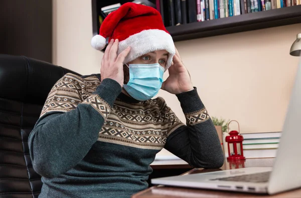 Noel online tebrikleri. Noel Baba şapkalı ve maskeli genç adam bilgisayar kullanarak arkadaşları ve çocukları arayarak şok etti ve şaşırttı. Coronavirus sırasında Noel.