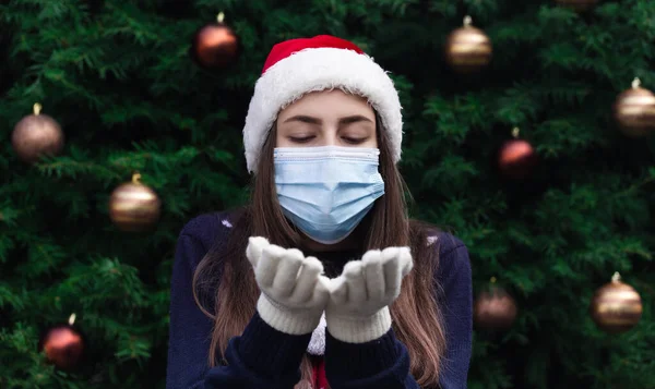 Christmas air kiss. Close up Portrait of woman wearing a santa claus hat and medical mask with emotion. Against the background of a Christmas tree. Coronavirus pandemic