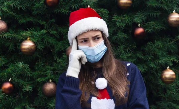 贈り物を選ぶ 閉じる感情を持つサンタの帽子と医療用マスクを身に着けている女性の肖像 クリスマスツリーを背景に コロナウイルスのパンデミック — ストック写真