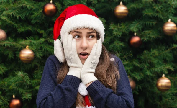 Shocked or surprised Christmas. Close up Portrait of woman wearing a santa claus hat with emotion. Against the background of a Christmas tree.