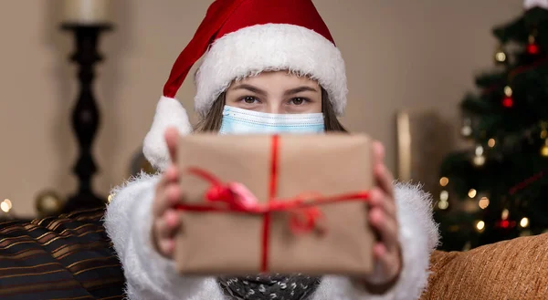 クリスマス マスクおめでとう 医学マスクでサンタの帽子と白いセーターを身に着けている肖像女性 赤いリボンとギフトプレゼントボックスを与えます 背景にクリスマスツリーのボケ — ストック写真
