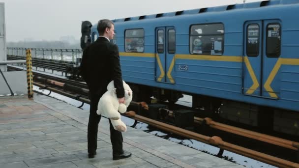駅でスーツ姿の青年が電車を待っている 手にはおもちゃの熊が — ストック動画