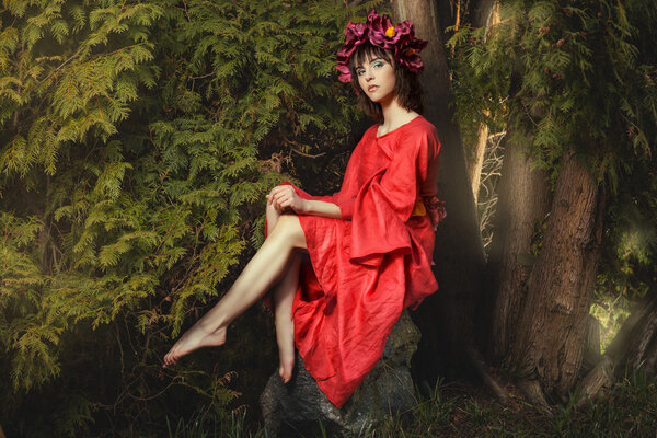 In the forest fairy sitting on a rock girl with red wreath of flowers on her head.