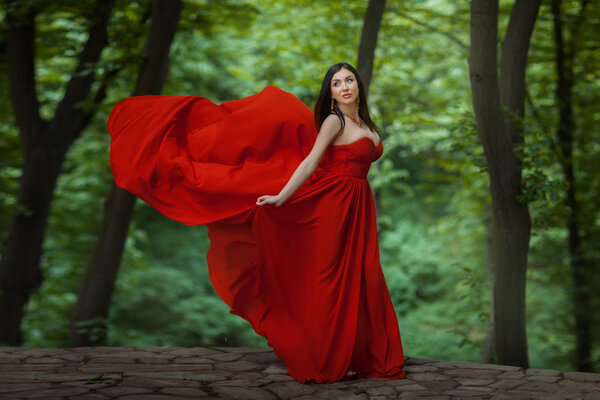 The girl is in the forest, her red dress develops wind. She stands on the precipice.
