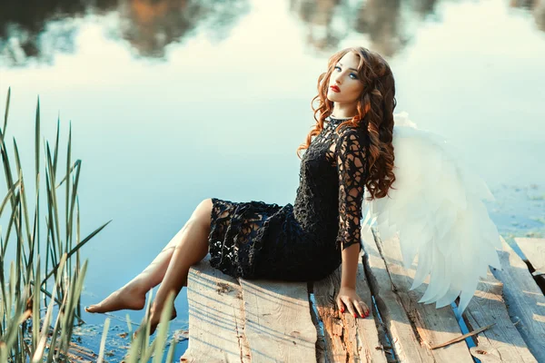 Girl with wings of an angel sitting near a river. — Stock Photo, Image