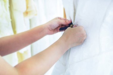 Kadın asistanın dükkânda gelinlik denemesine yardım ettiği eli kapat. Asyalı genç kadınlar gelinlik alışverişi yapıyor, askılarda bir sürü beyaz elbise asılı.. 