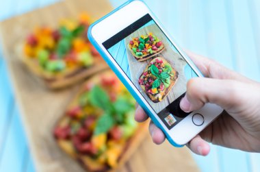 Hands taking photo with smartphone of bruschetta on wooden cutting board