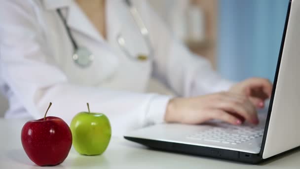 Doctora escribiendo en el teclado del portátil, comiendo una manzana. Forma de vida saludable — Vídeos de Stock