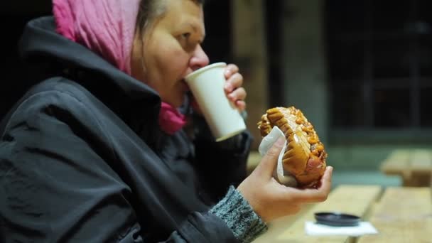 Голодная взрослая женщина пьет чай и ест хот-доги на благотворительном мероприятии, фаст-фуд — стоковое видео