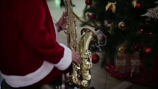 Saxofonist i Santa Claus Suit spela Merry Christmas Carol nära New Year Tree — Stockvideo