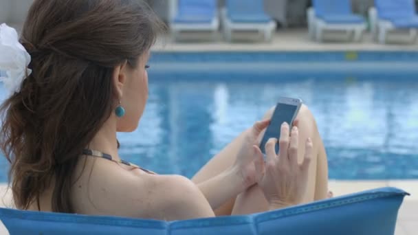 Красивая женщина печатает сообщения, смс-ки на смартфоне возле бассейна — стоковое видео