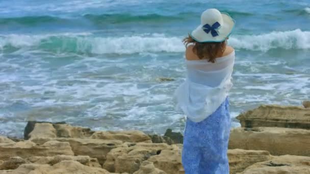 Lonely female wrapped in scarf watching foamy ocean waves crash on rocky beach — Stock Video